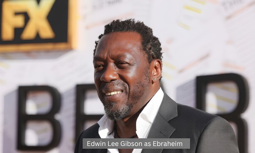 Edwin Lee Gibson attends The Bear premiere event, posing in a dark suit in front of the show's promotional backdrop.
